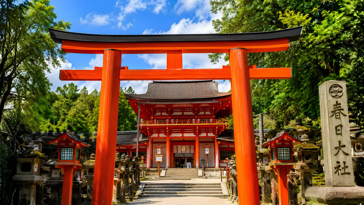 Kasuga Taisha Shrine
A vibrant red Japanese torii gate stands prominently in front of a traditional red shrine building, surrounded by lush green trees under a blue sky with white clouds. A stone monument with Japanese characters is visible on the right side.